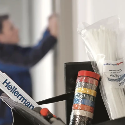 A toolbox in the foreground featuring HellermannTyton cable ties, a stack of colorful electrical tape, and the edge of a ruler. In the blurred background, a craftsman in blue workwear is working on a wall.