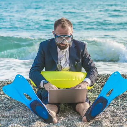 Ein Mann sitzt im Anzug mit Schnorchelmaske, Schwimmflossen und Schwimmring am Strand vor einem Laptop. Hinter ihm rauschen die Wellen. Die Szene wirkt humorvoll und kontrastreich zwischen Büroarbeit und Strandurlaub.
