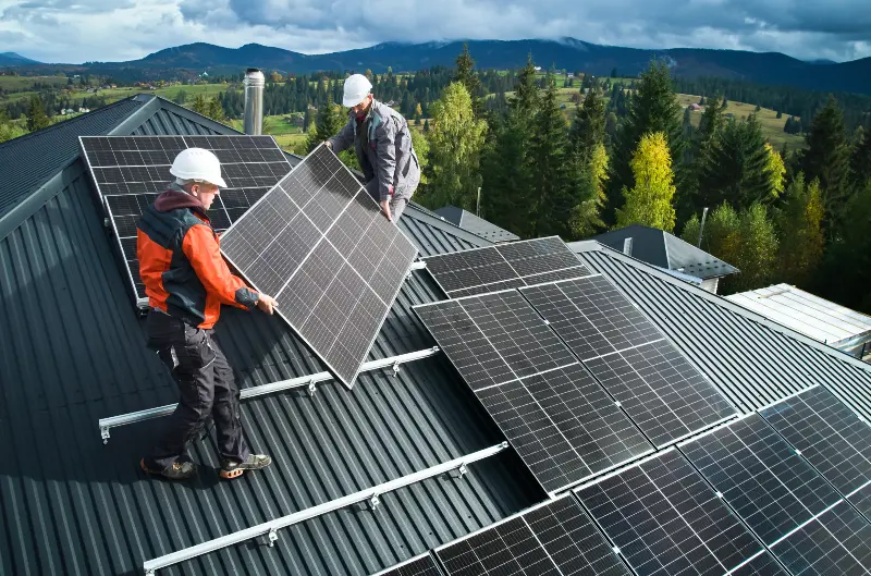 Two workers install solar panels on a sloped metal roof with a mountainous landscape in the background.