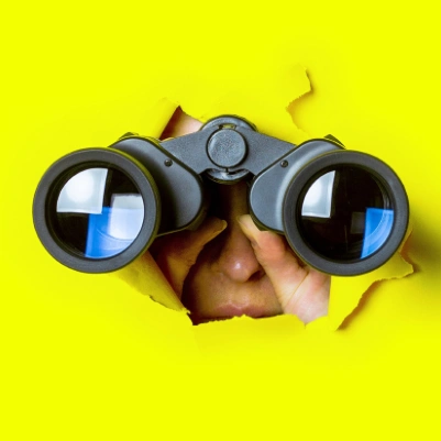 Person looking through binoculars through a hole torn in yellow paper.