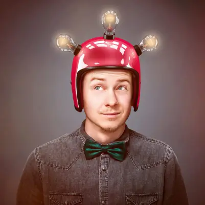 Eine Person trägt ein graues Jeanshemd mit grüner Fliege und einen roten Helm, auf dem drei leuchtende Glühbirnen wie Antennen befestigt sind. Das Gesicht der Person ist unscharf dargestellt. Der Hintergrund ist neutral und leicht abgedunkelt.