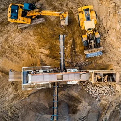 Luftaufnahme einer Baustelle mit zwei gelben Baggern, Förderanlage und Materialhaufen auf sandigem Untergrund.