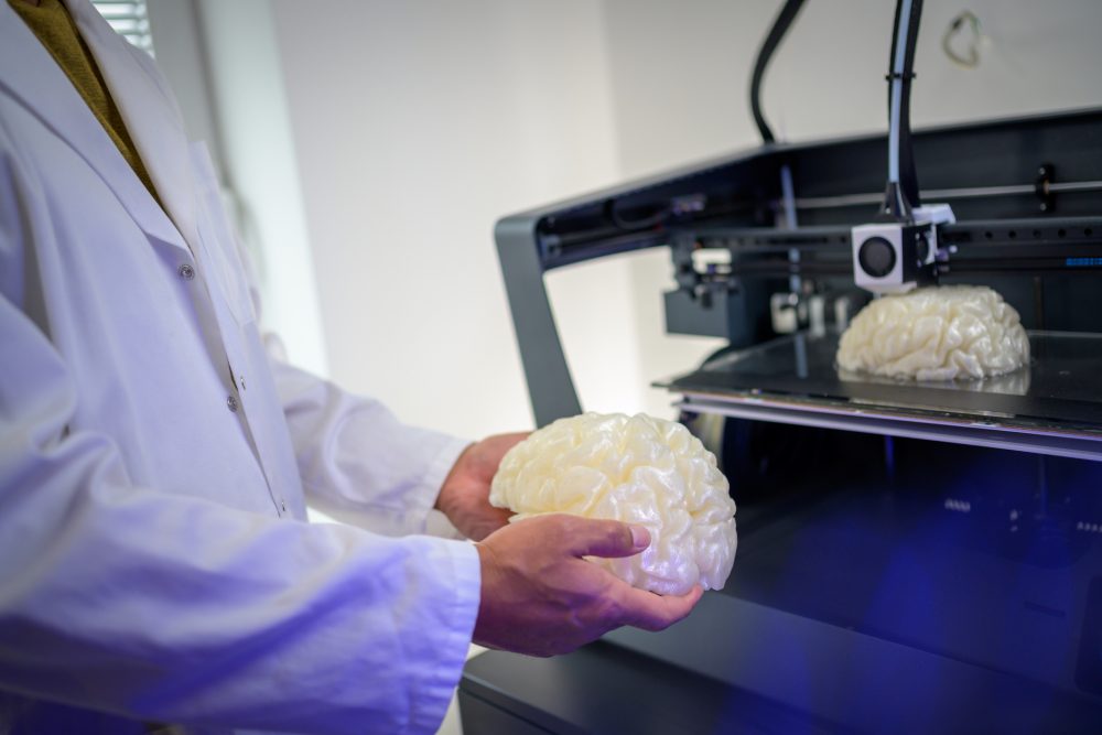 3D printing in Medicine - Male hands holding human brain model. Next item is in 3D printer. Producing of 3D object from digital model using additive manufacturing.