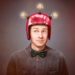 Mann mit einem rotem Helm, der drei Glühbirnen aufgebracht hat. Der Mann trägt Hemd und Fliege.